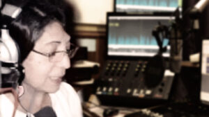 Kuva radio-ohjelmaa tekevästä naisesta, henkilö kuvan vasemmassa reunassa, taka-alalla näkyy radiostudiota monitoreineen ja mikrofoneineen.