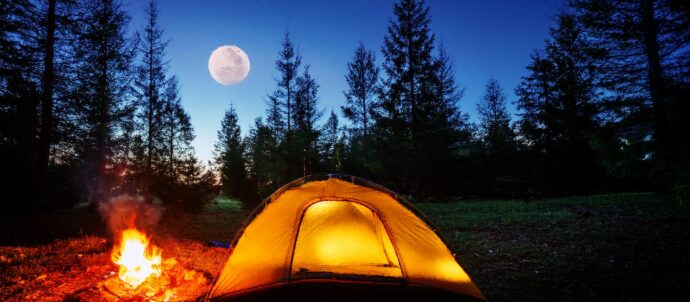 Tekoälyllä luotu valokuva öisestä metsästä, jossa täysikuu kirkkaalla taivaalla, etualalla keltainen teltta ja nuotio sen vieressä.