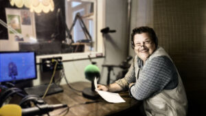 Toimittaja istuu pöydän ääressä radiostudiossa paperi kädessä, katsoen kameraan.