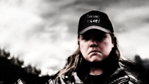 Rasteroitu, lähes harmaasävyinen kuva toimittajasta, jolla musta lippalakki, pitkät hiukset ja farkkuliivi.