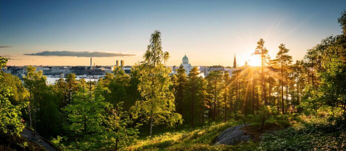Tekoälyllä luotu kuva kuvitteellisesta Helsingistä, jossa aurinko laskee kaupunkisiluetin taakse ja etualalla on metsää.