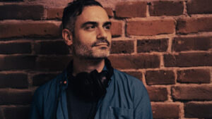 Mies seisoo punaisen tiiliseinän edessä katsoen mietteliäästi vasemmalle yläviistoon.