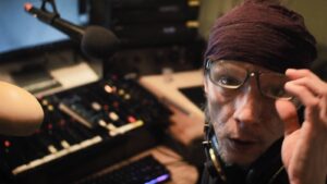 Äänipöydän ääressä oleva toimittaja katsoo suoraan kameraan, pitäen kiinni silmälaseistaan.