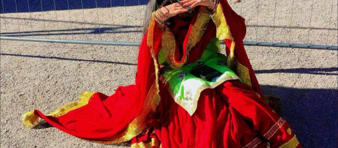 Aurinkolasipäinen, tummahiuksinen nuori nainen värikkäässä mekossa istuu verkkoaidan edessä.